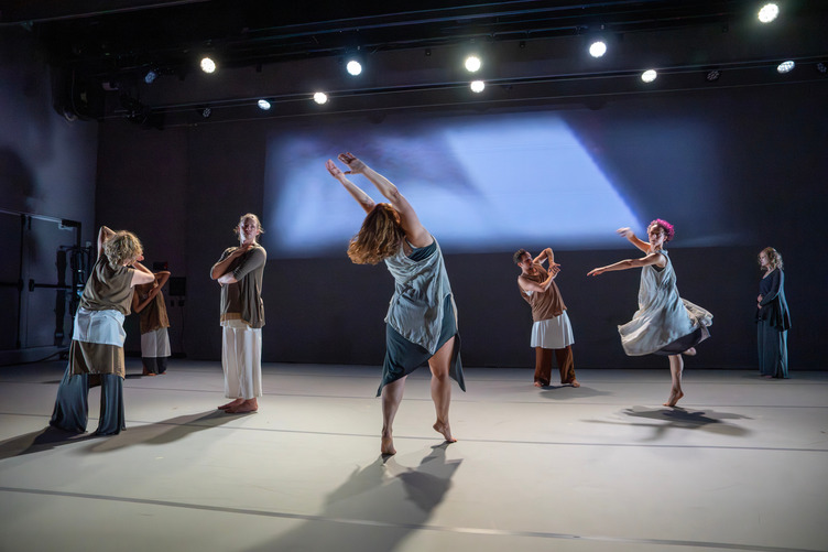 Seven dancers in various standing positions, including one long-haired woman dancer on releve with arms extended, as another dancer with short, red hair is caught turning in passe.